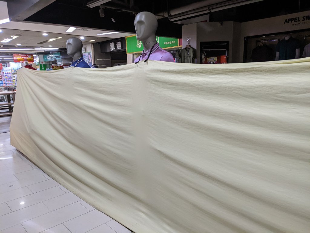 Blanket suspended from mannequins protecting a clothes store with my grocery store in the background.